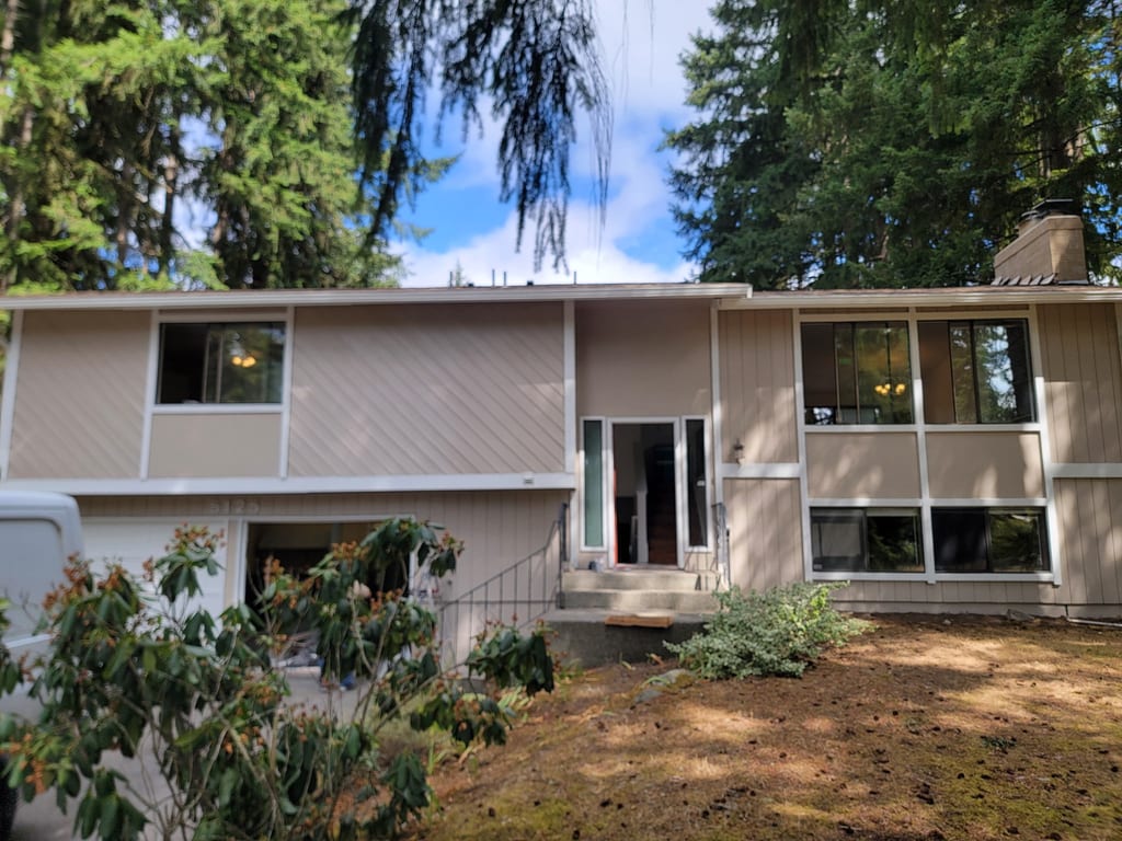 Split-level home under Douglas firs with the roofline stripped and prepped for new seamless gutters.